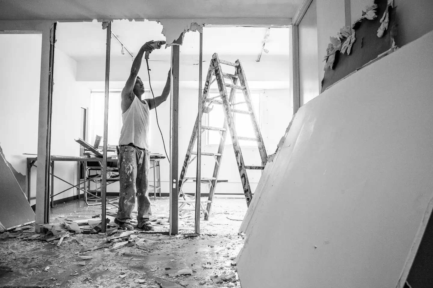 Ouvrier sur un escabeau qui fixe une ossature au plafond, en noir et blanc