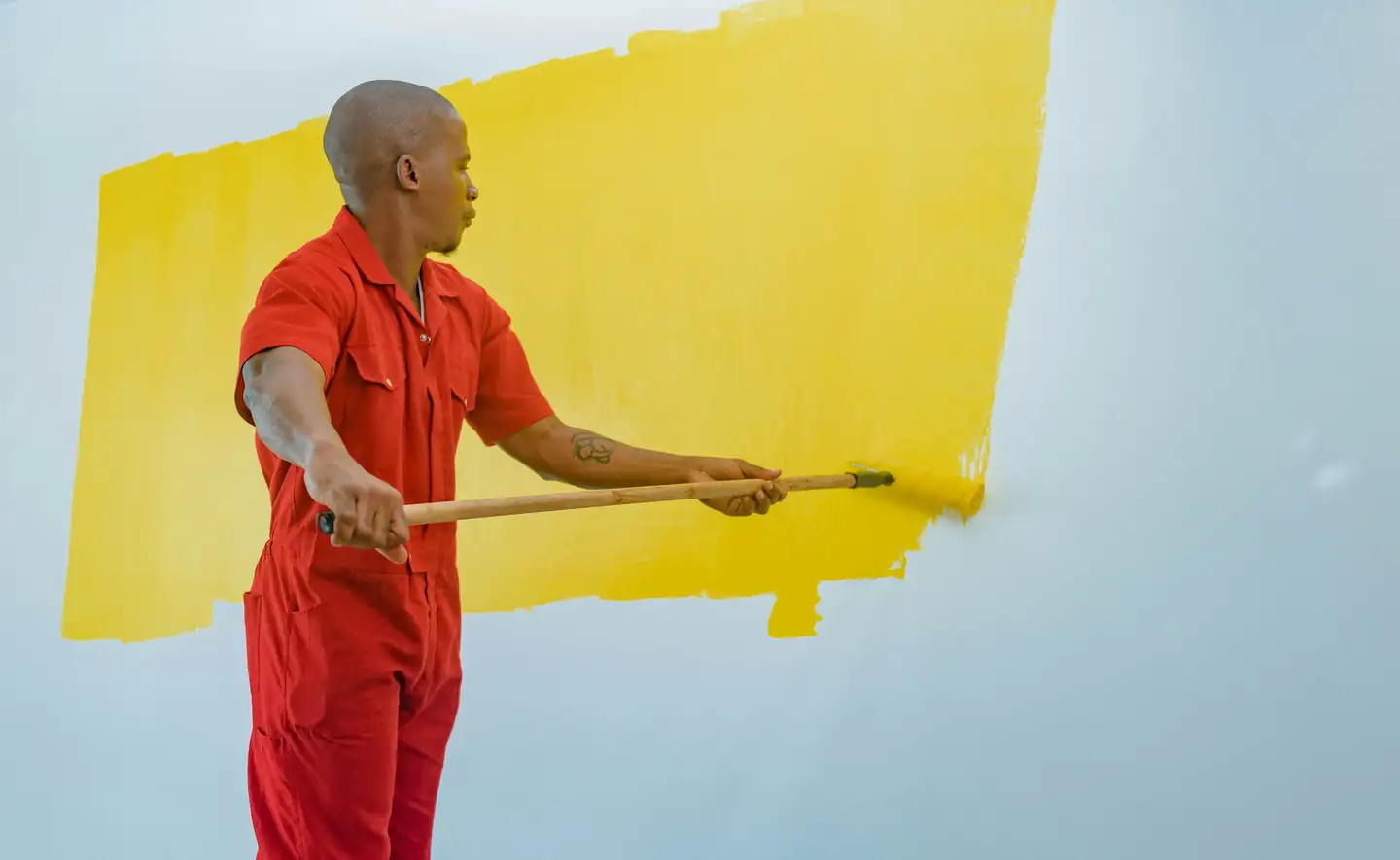 Peintre en combinaison rouge qui passe un rouleau de peinture jaune sur un mur clair